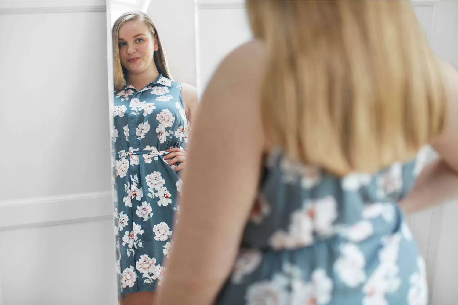 Woman in blue floral dress looking at her body image in the reflection of a mirror.