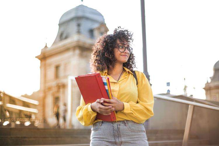 College student wearing a yellow shirt holding books on campus.