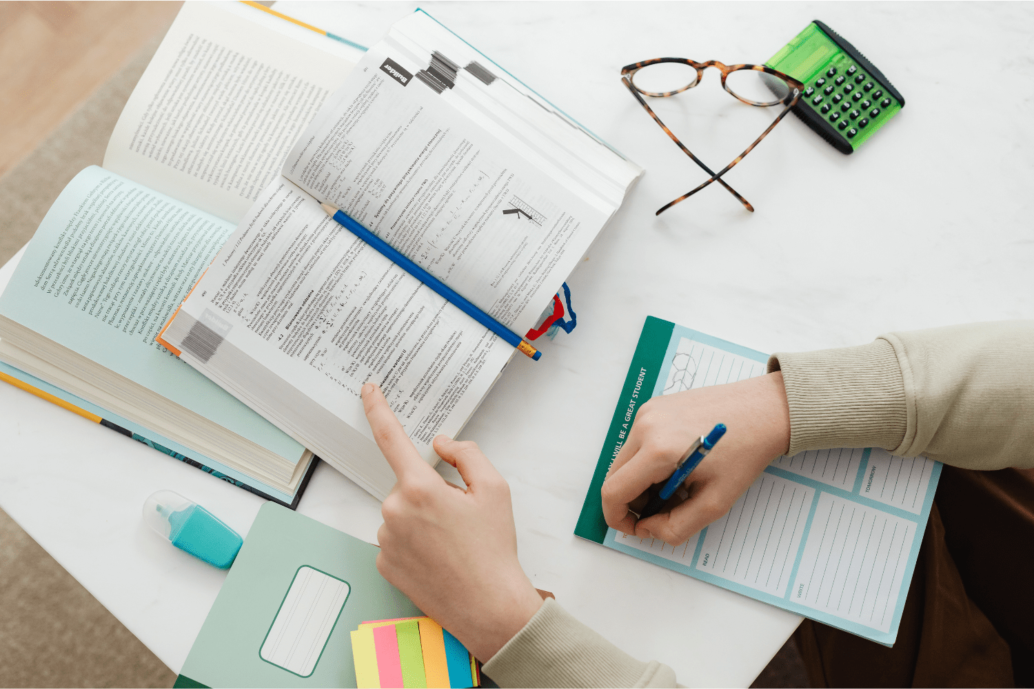 A student studying at a desk with open textbooks, a to-do list notepad, colorful sticky notes, and a calculator. Glasses rest nearby, suggesting intense focus during exam preparation.