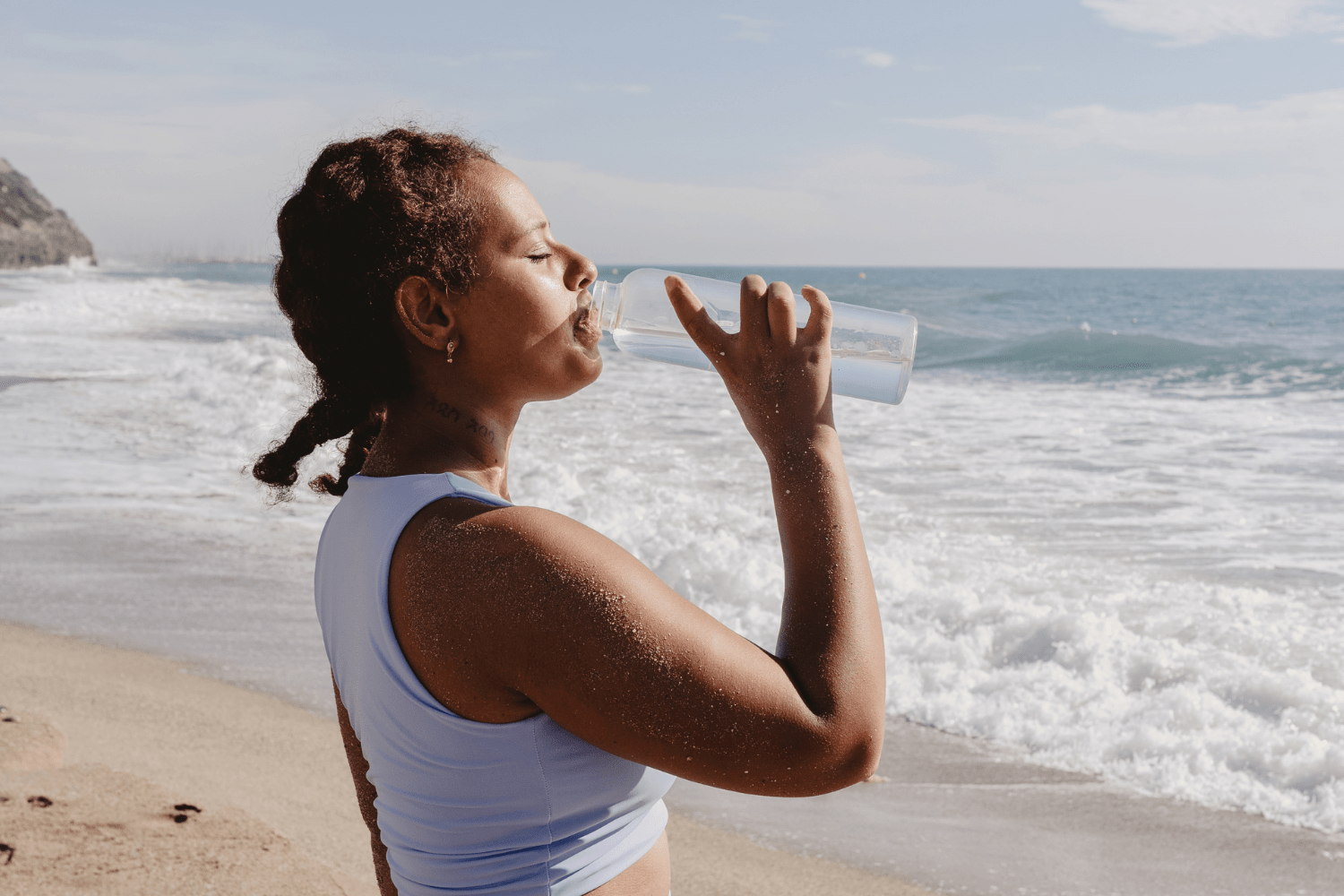 Person drinking water from a clear bottle while standing on a sandy beach with ocean waves in the background, emphasizing the importance of staying hydrated during summer.
