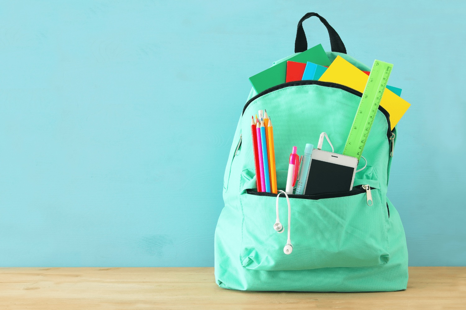 A turquoise backpack filled with school supplies including pencils, pens, notebooks, a ruler, and a smartphone, against a light blue background.