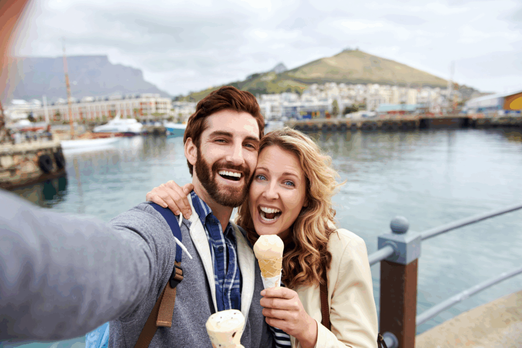 A smiling couple taking a selfie outdoors near a waterfront, holding ice cream cones, with mountains and buildings in the background, illustrating managing travel food anxiety.