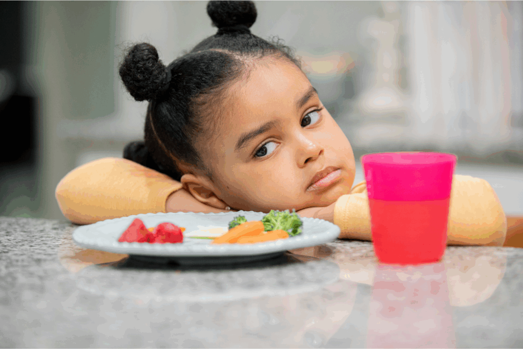 A young girl with her head resting on her arms, looking away thoughtfully at a table with a plate of strawberries, carrots, broccoli, and a pink cup.