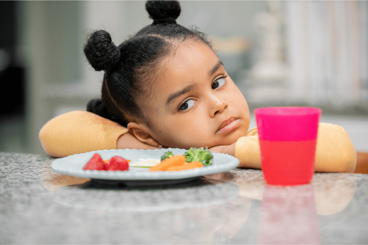 A young girl with her head resting on her arms, looking away thoughtfully at a table with a plate of strawberries, carrots, broccoli, and a pink cup.