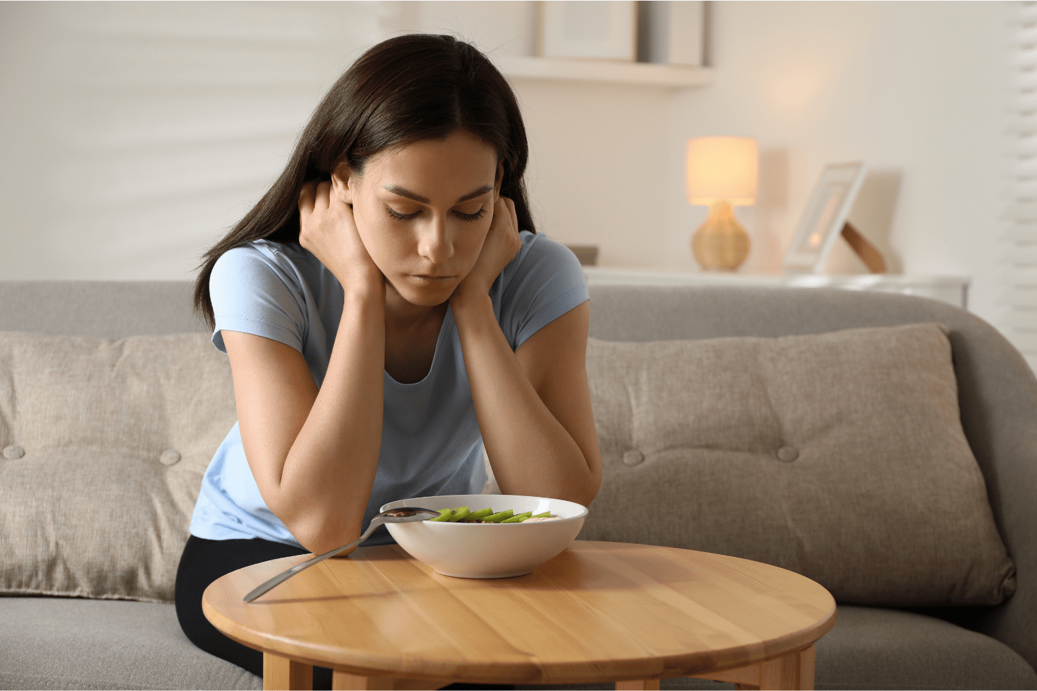 A young woman sitting on a couch, looking down anxiously at a bowl of food, illustrating the emotional struggle behind eating disorders and challenging the ‘Myth of Willpower.’